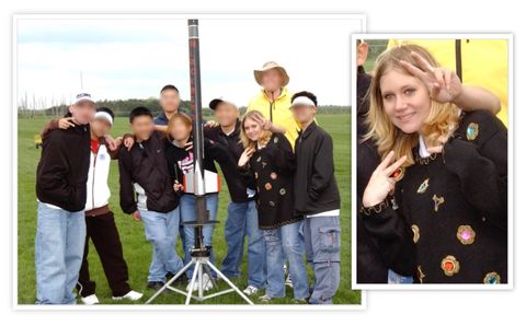 A group of nine people are posing for a picture in an open field. Directly in front of them in the center of the image is a tall, thin rocket. Dallas Humber is the second from the right. She’s wearing blue jeans and a black sweater adorned with gold appliques and oversized multi-colored jewels. She has shoulder length blonde hair and is making a double handed “victory” gesture.